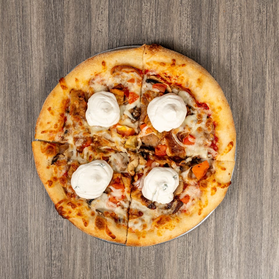 Overhead view of a loaded cheese pizza topped with vegetables, mushrooms, meat, and dollops of white cream on a wooden table.
