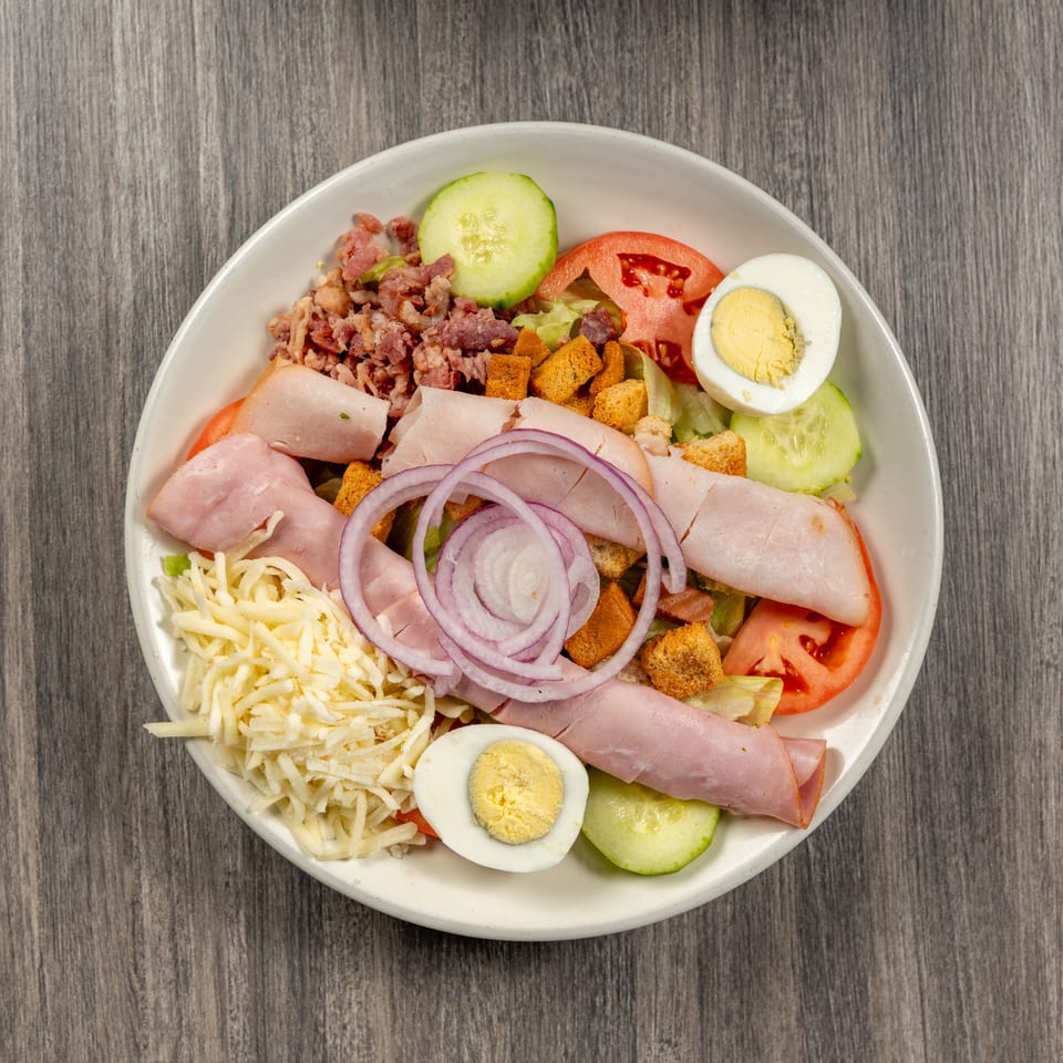 Overhead view of a loaded salad with ham, bacon, boiled eggs, cheese, cucumbers, tomatoes, onions, lettuce, and croutons.