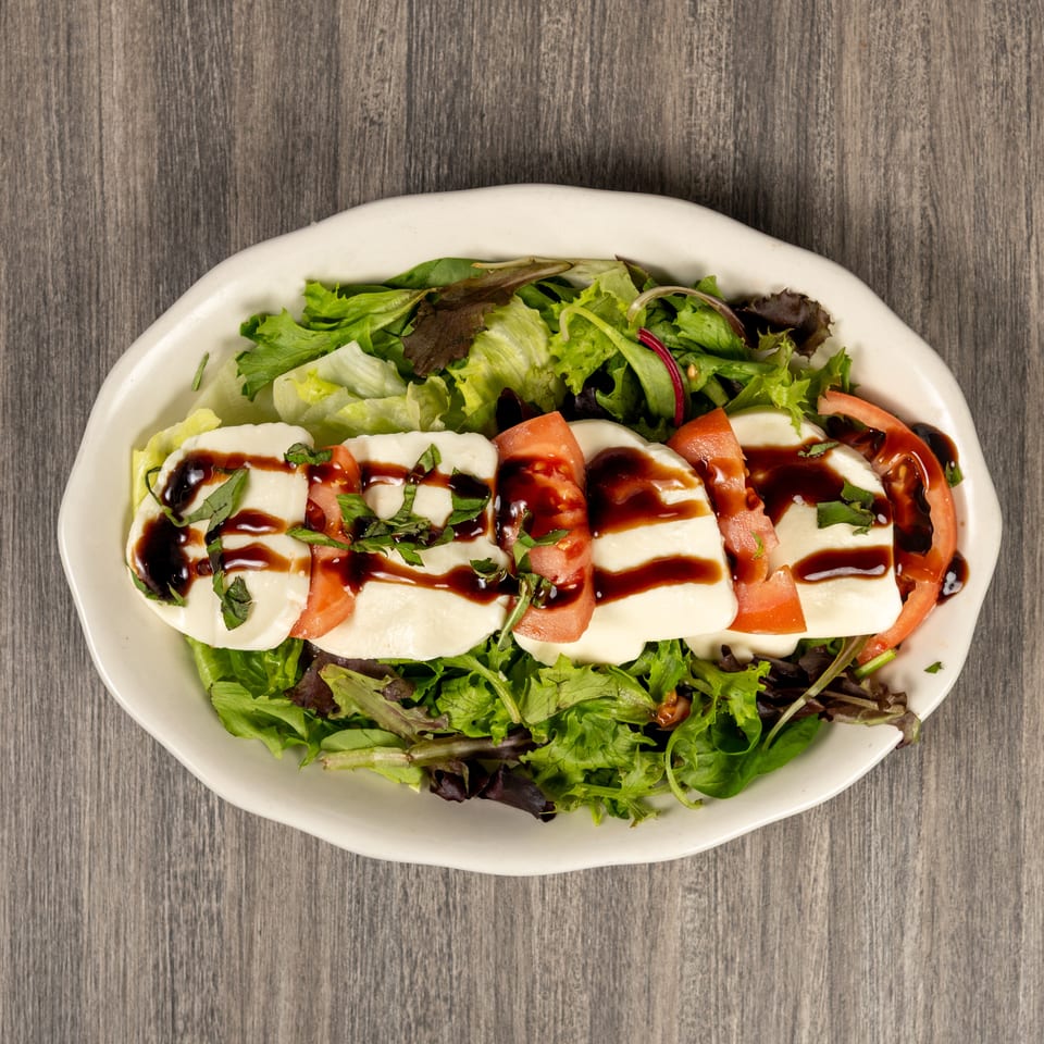 Mixed green salad topped with sliced mozzarella, tomatoes, fresh herbs, and balsamic glaze on a white oval plate.