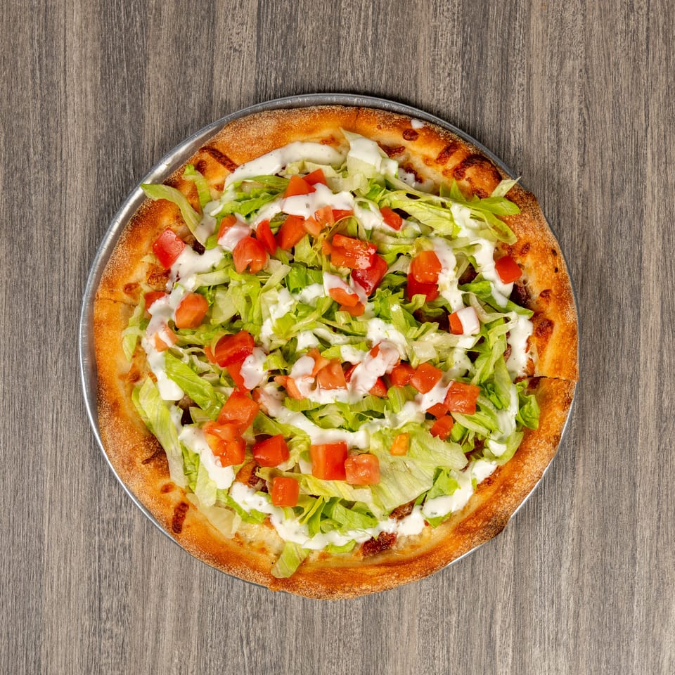 Overhead view of a baked pizza topped with shredded lettuce, diced tomatoes, and creamy white dressing on a metal pan.
