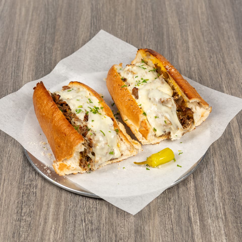 Two cheesesteak sandwiches with melted cheese, mushrooms, and parsley on a paper-lined plate with a pepperoncini.