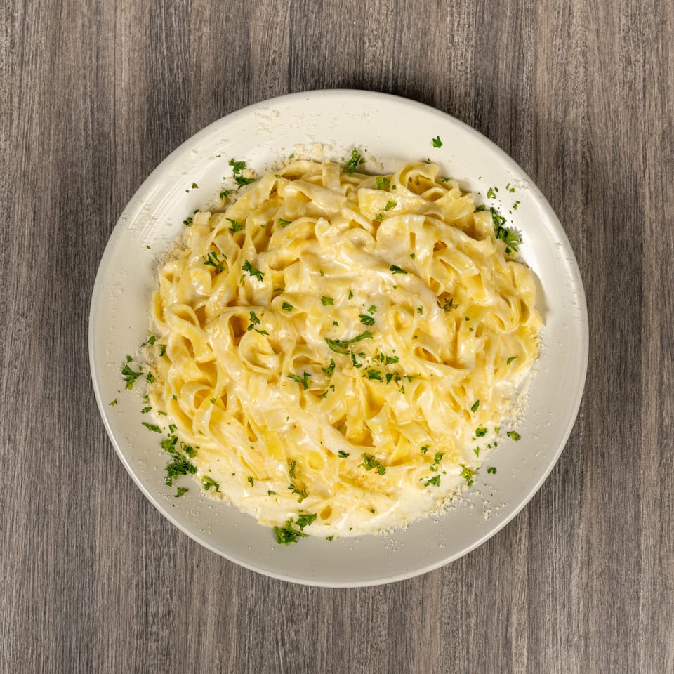 Creamy fettuccine Alfredo topped with parsley and grated Parmesan on a white plate atop a wood-grain table.