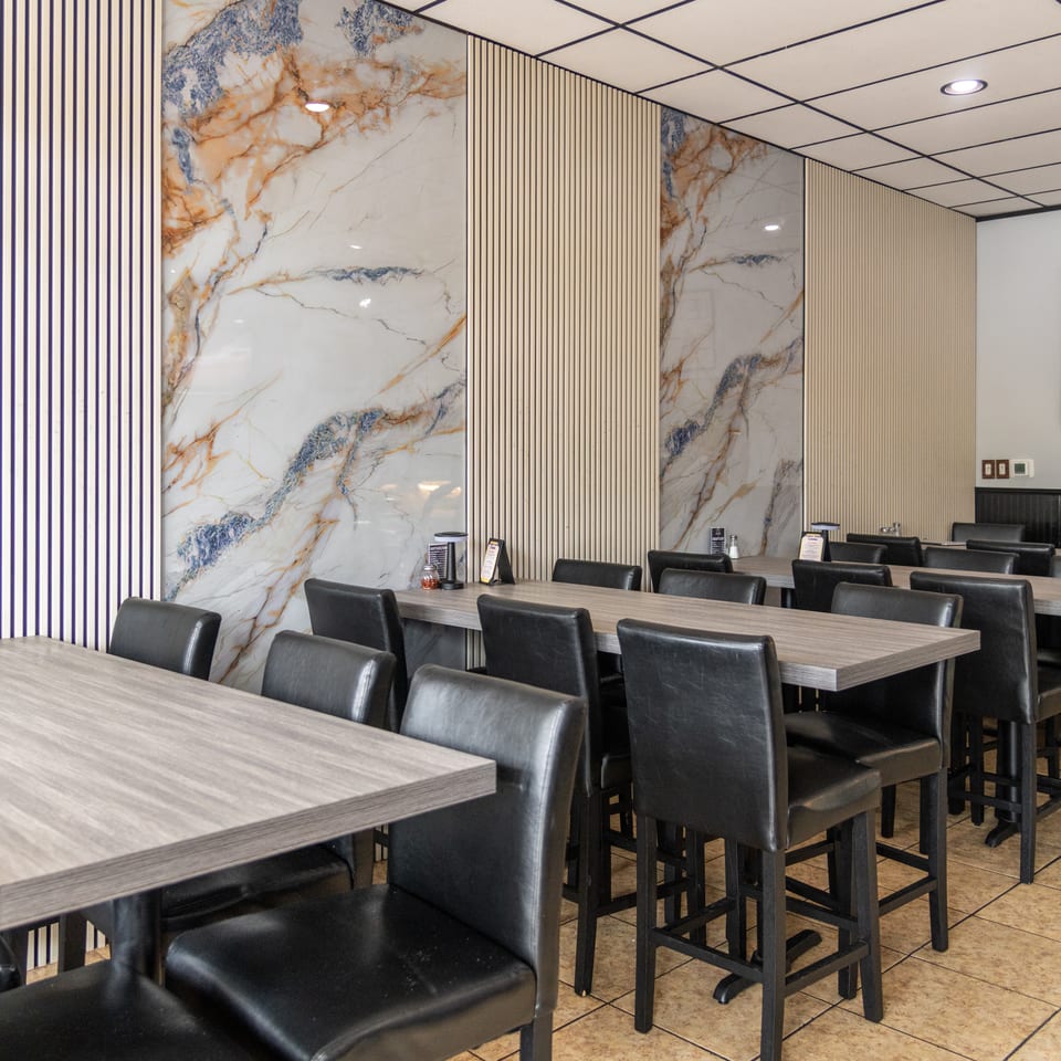 Modern restaurant dining area with gray tables, black leather chairs, striped walls, marble accents, and a Rat Pack poster.