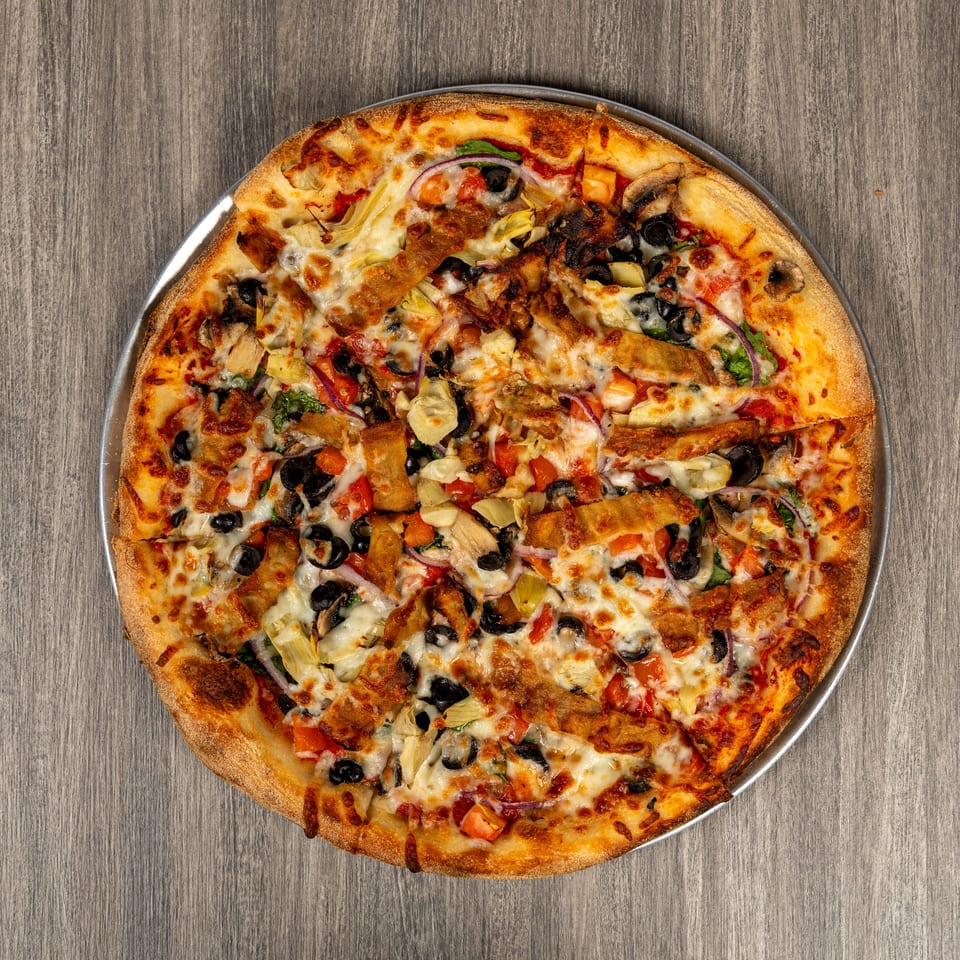 Overhead view of a loaded pizza with melted cheese, black olives, mushrooms, artichokes, tomatoes, onions, and crispy chicken on a metal tray.