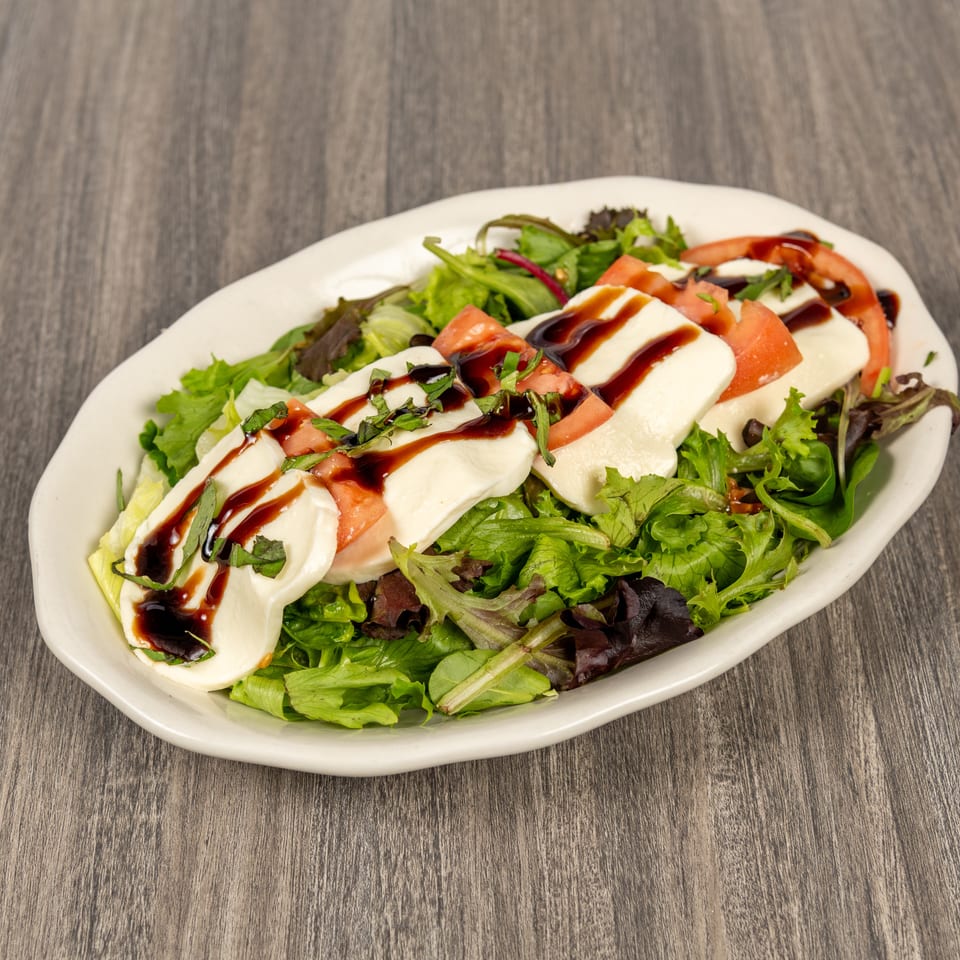 Mixed green salad topped with sliced mozzarella, tomatoes, basil, and balsamic glaze on a white plate.