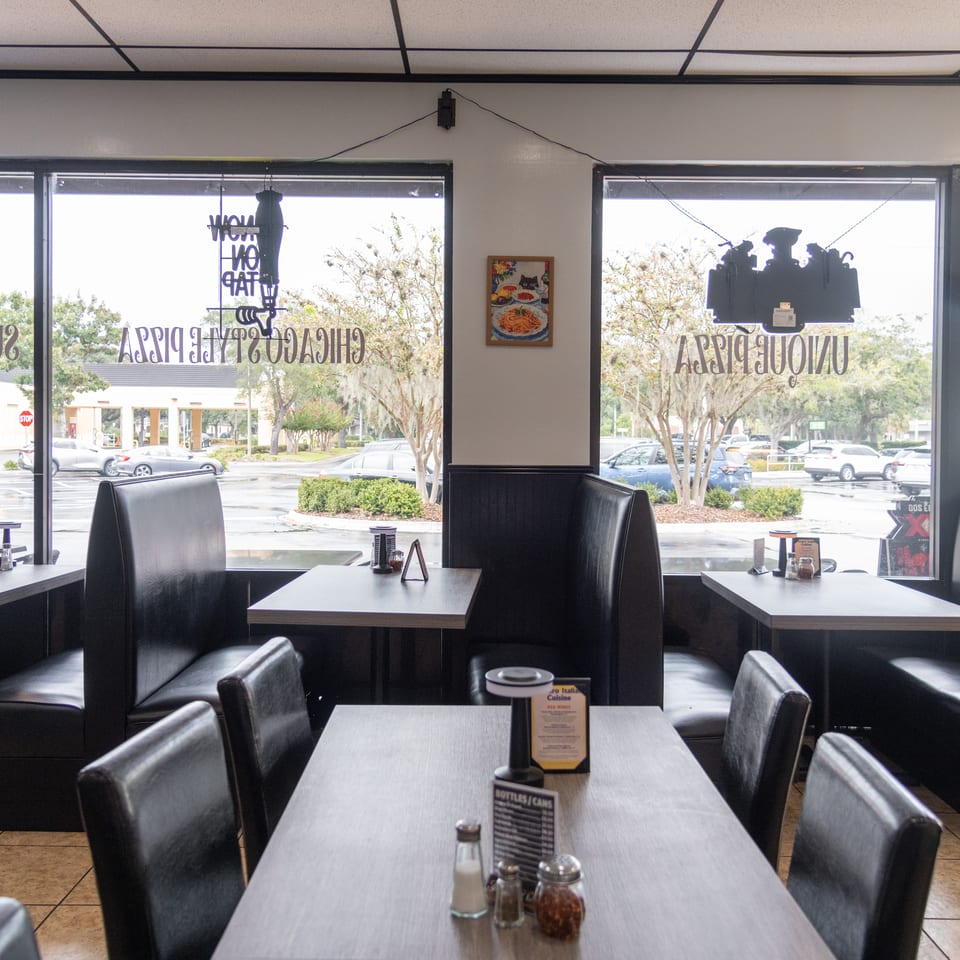 Bright Italian restaurant dining room with black leather booths, neatly set tables, and large windows overlooking a parking lot.