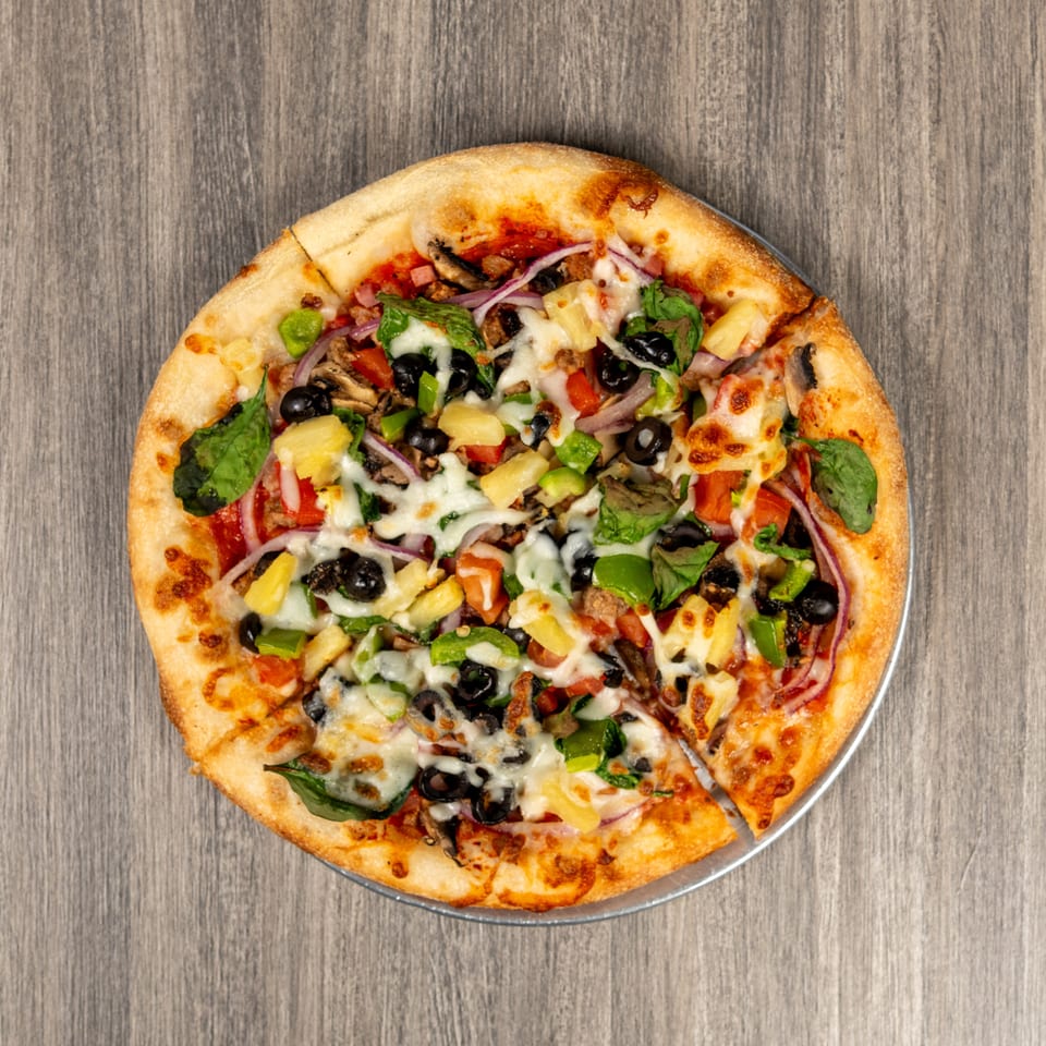 Overhead view of a loaded pizza topped with melted cheese, pineapple, black olives, peppers, mushrooms, onions, spinach, and tomatoes.