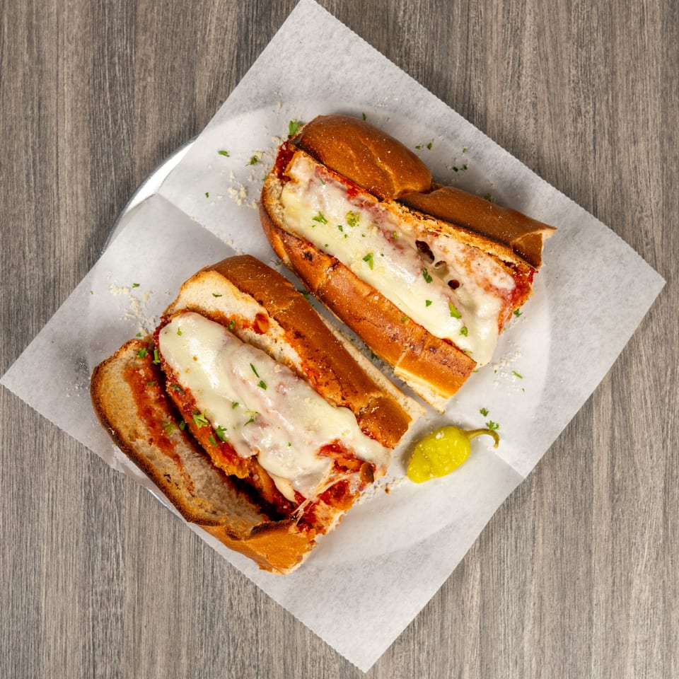 Two toasted meatball subs topped with melted cheese and parsley, served on parchment with a pepperoncini.