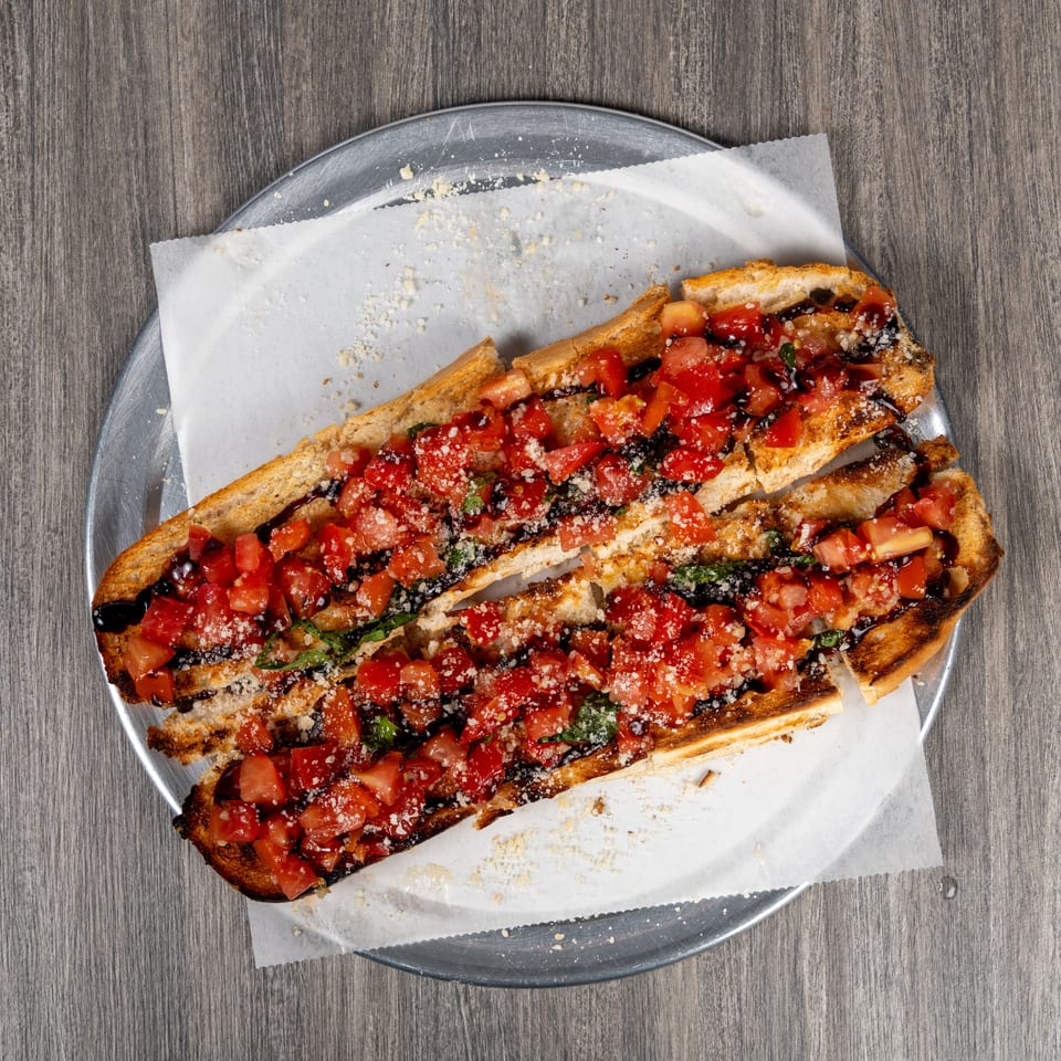 Grilled bruschetta topped with diced tomatoes, basil, balsamic glaze, and grated cheese on a metal plate.