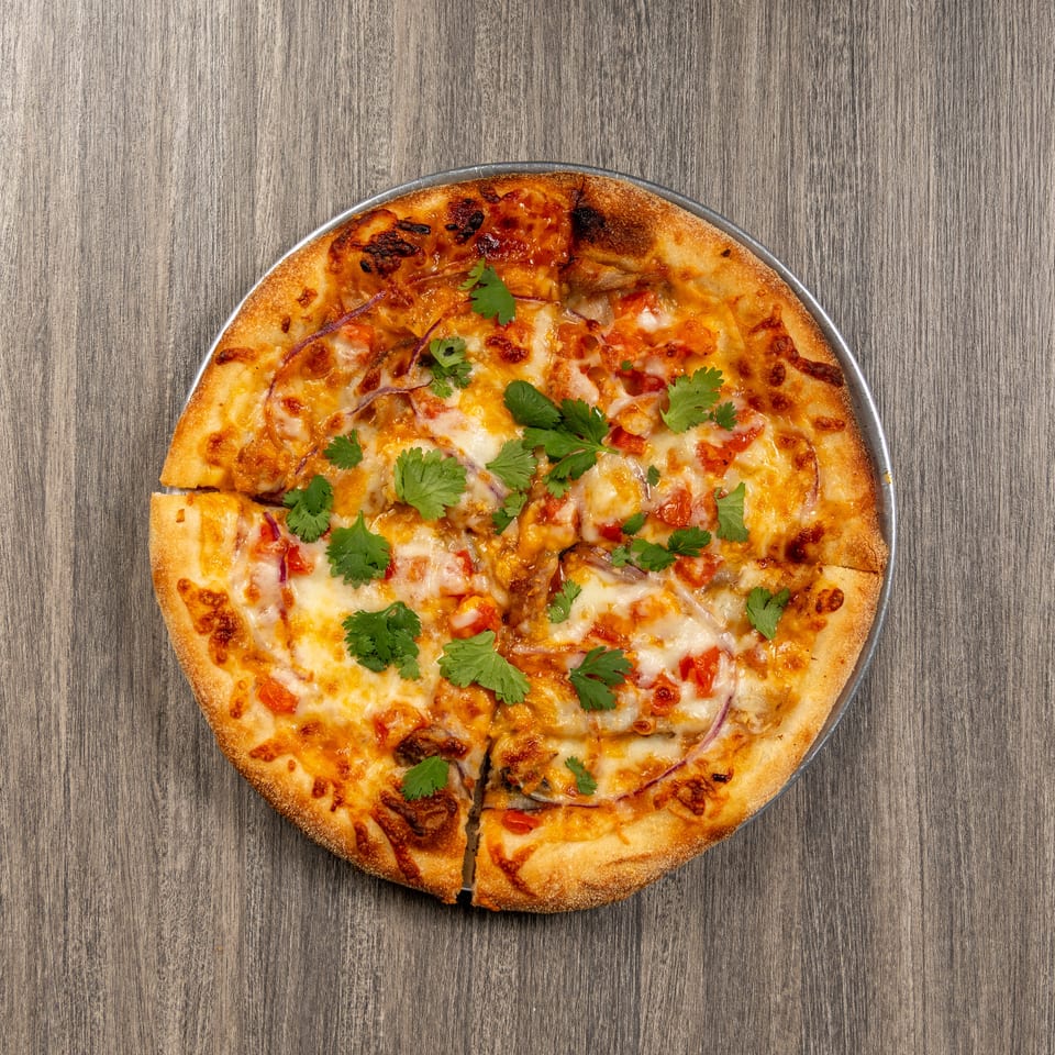Cheesy pizza topped with tomatoes, red onions, roasted vegetables, and fresh cilantro on a metal tray over a wooden table.