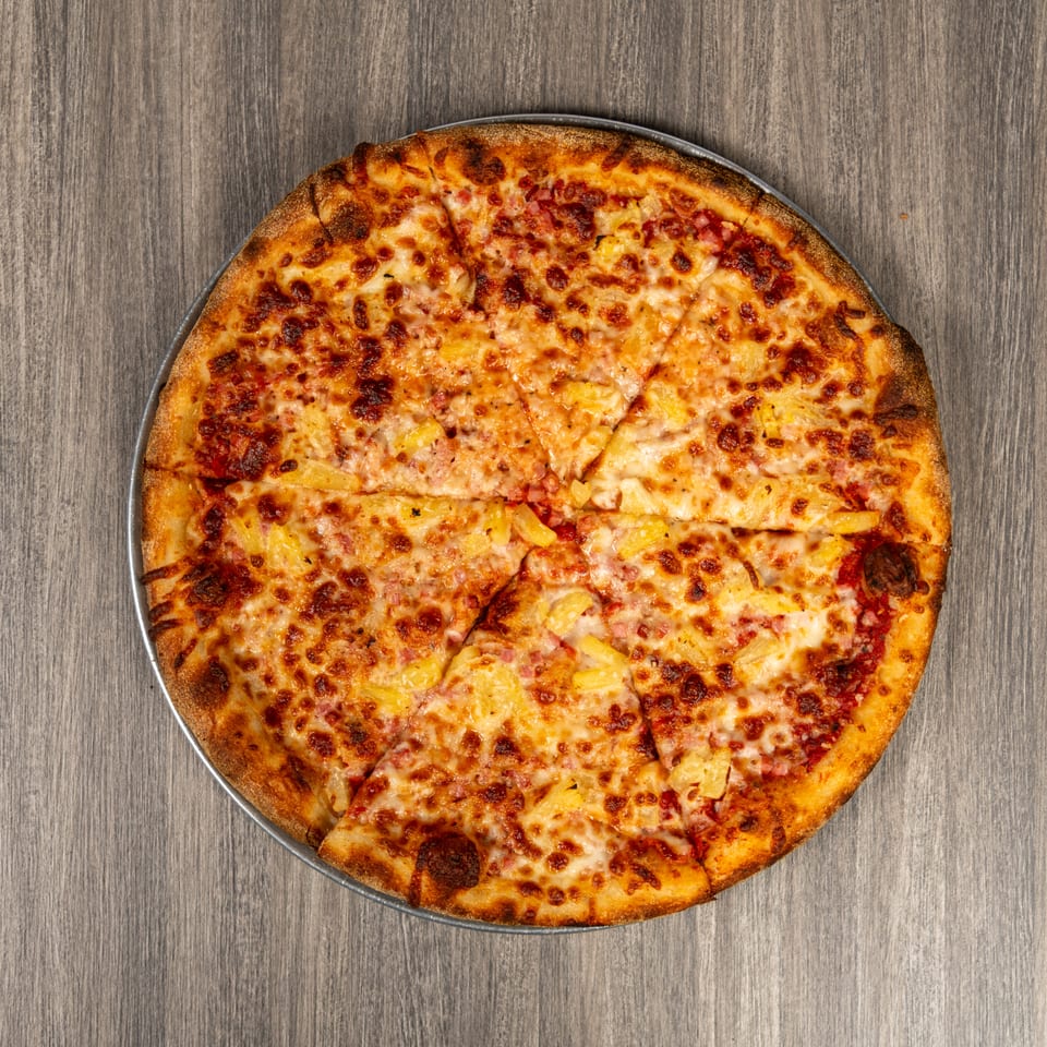 Large cheese pizza topped with ham and pineapple, sliced and served on a metal pan over a wood-grain table.